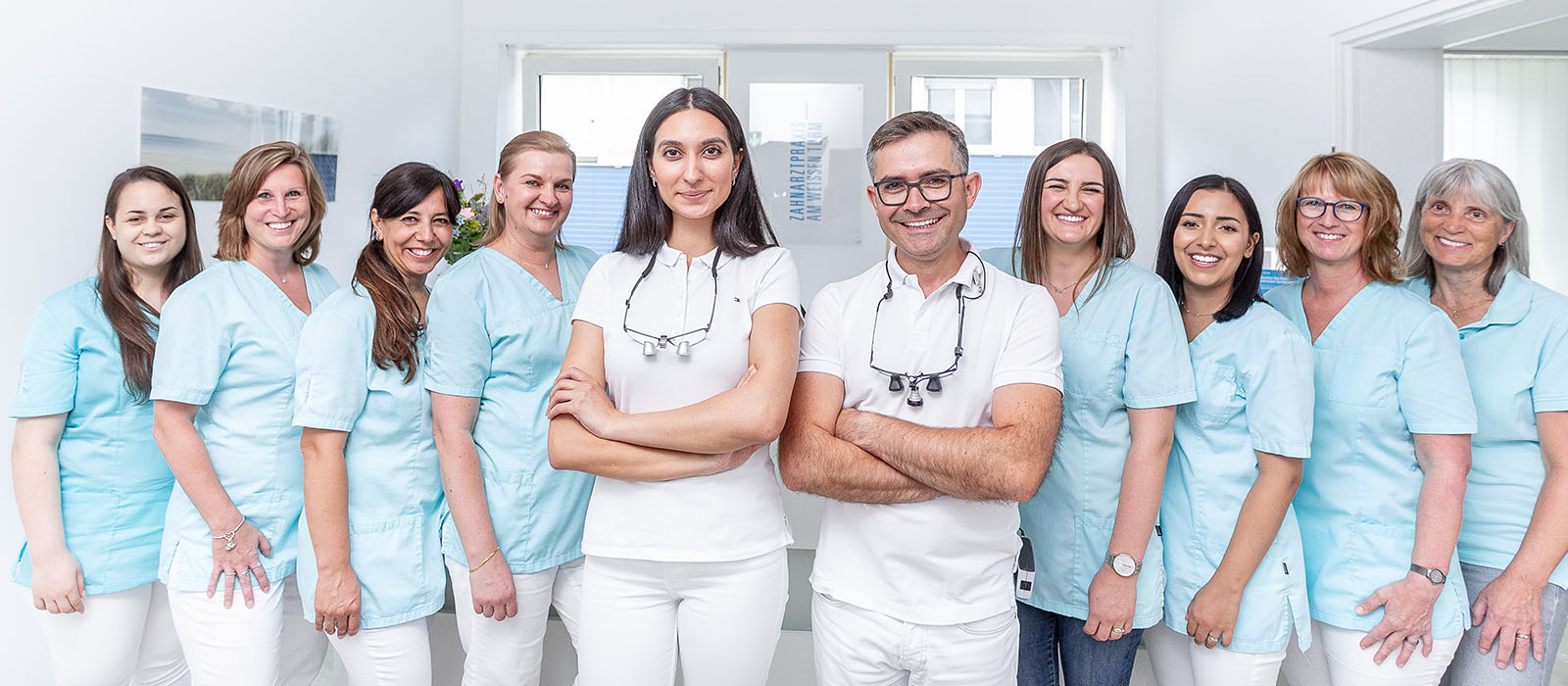 Das Team von Zahnarzt Daniel Krutsch mit Ihm und Zahnärztin Celine Nazzal inweißer Kleidung in der Mitte, die anderen Mitglieder reihen sich links und rechts daneben harmonisch zum Gruppenfoto zusammen.